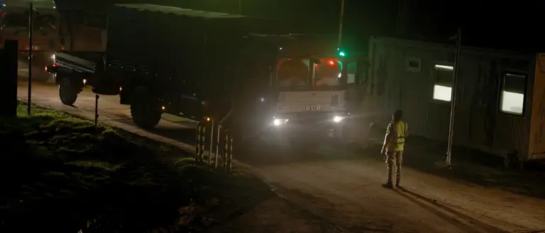 Large army transport vehicle being guided by a soldier in camouflage uniform at night.