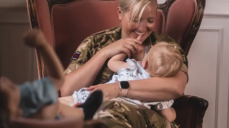 A woman in army uniform sits on a plush chair, smiling warmly at a baby on her lap. The setting is cozy, with a sense of tenderness and joy.
