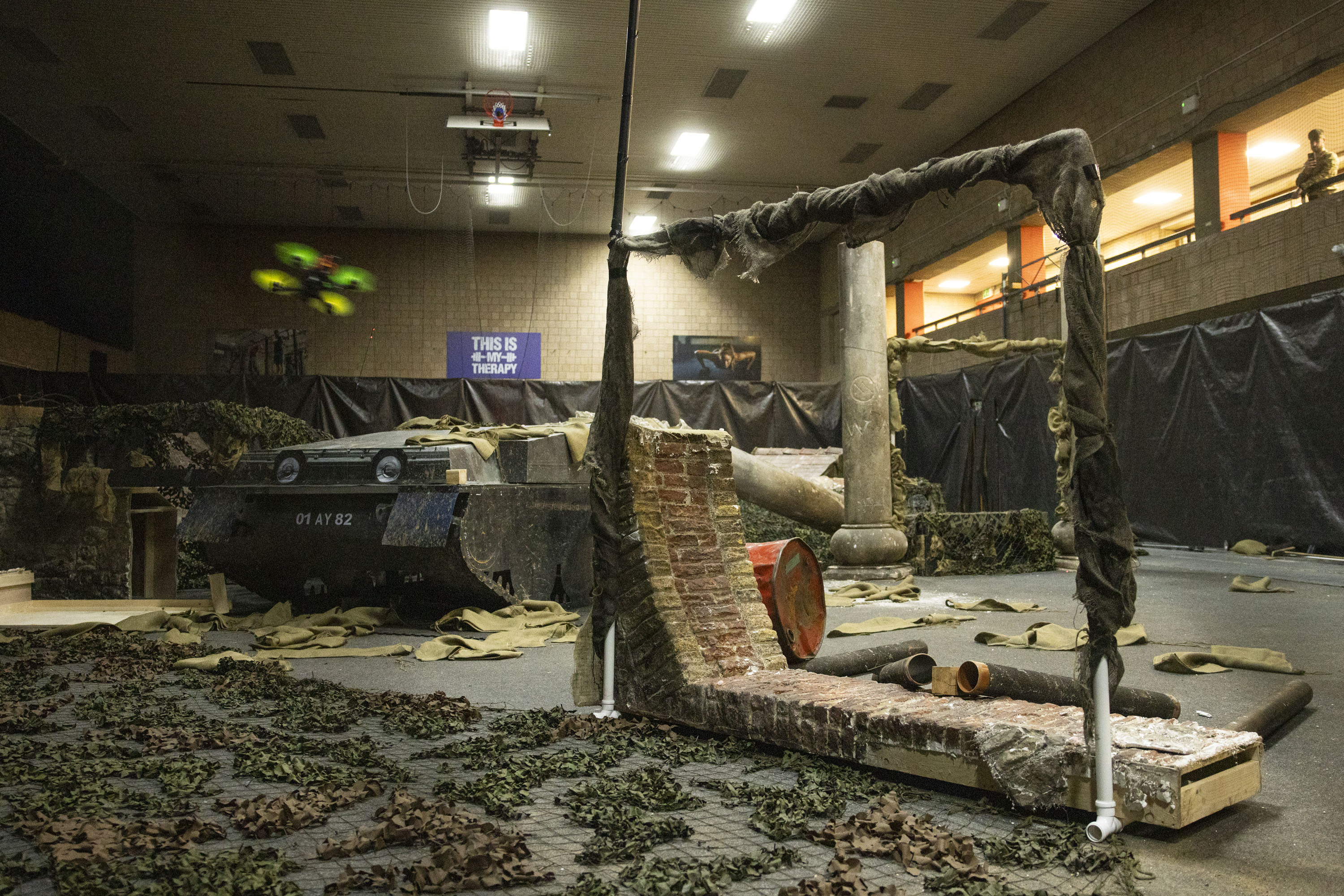 A drone flies through an artificially made war zone, a military vehicle is pictured in the background.