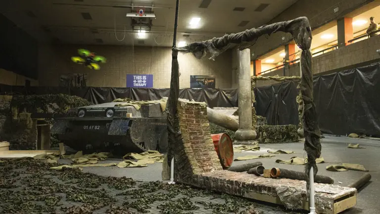 A drone flies through an artificially made war zone, a military vehicle is pictured in the background.
