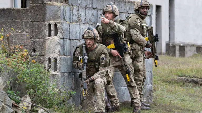 Three soldiers in camouflage gear and helmets stand alert by a brick wall. One kneels and looks ahead while another points. They all hold rifles.