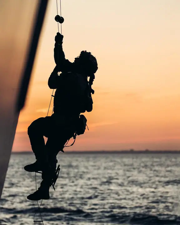 A person ascends a rope against the backdrop of a sea at sunset. The sky is a gradient of orange and pink.