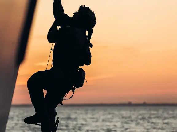 A person ascends a rope against the backdrop of a sea at sunset. The sky is a gradient of orange and pink.