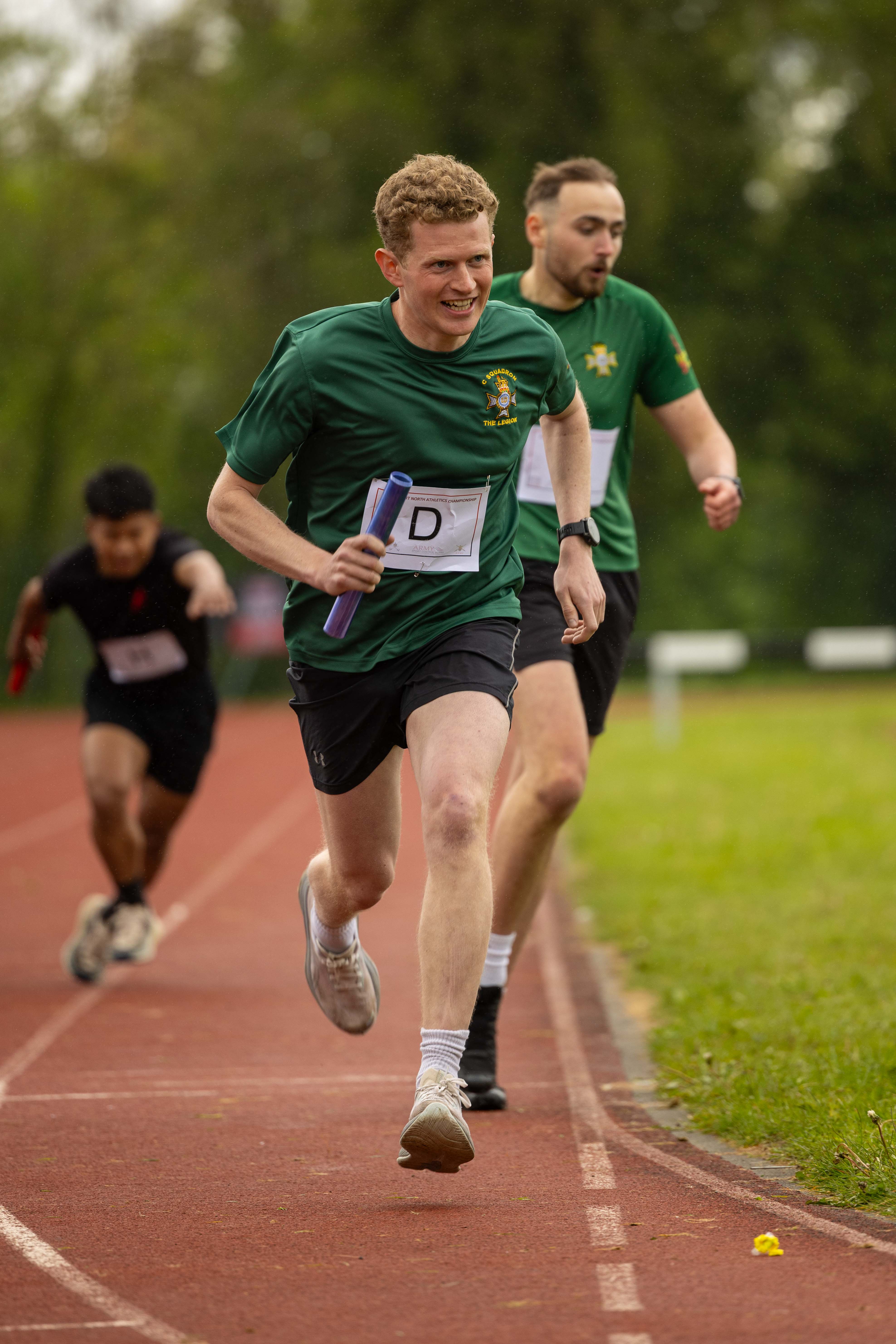 A person in a green shirt and shorts runs on a track, holding a baton. Two other individuals follow behind. 