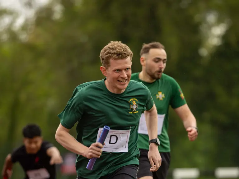 A person in a green shirt and shorts runs on a track, holding a baton. Two other individuals follow behind.