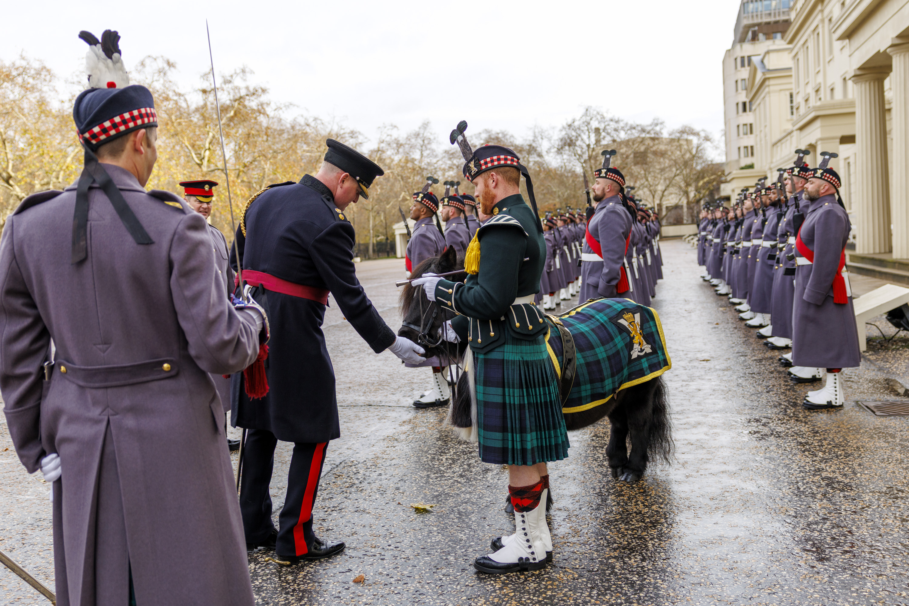 A military officer in a dark uniform stands in front of a soldier in a green kilt and traditional Scottish attire.