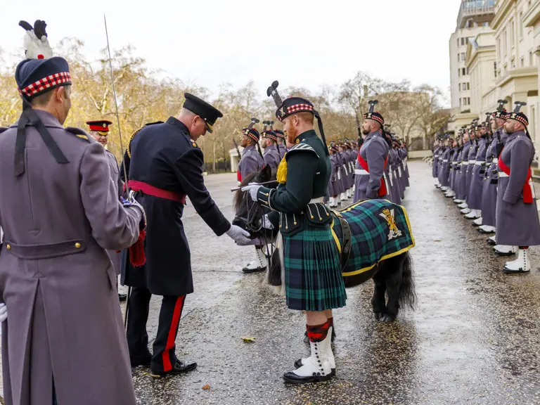 A military officer in a dark uniform stands in front of a soldier in a green kilt and traditional Scottish attire.
