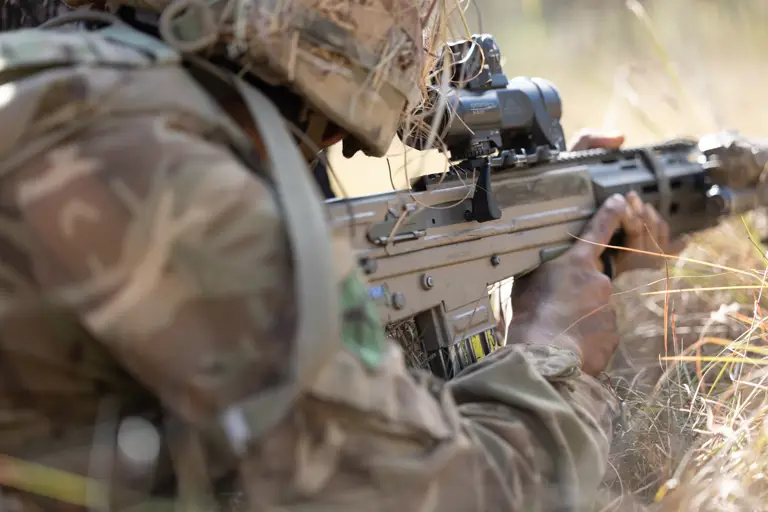  Soldier in camouflage gear aiming a scoped rifle while lying prone in tall grass during daylight.