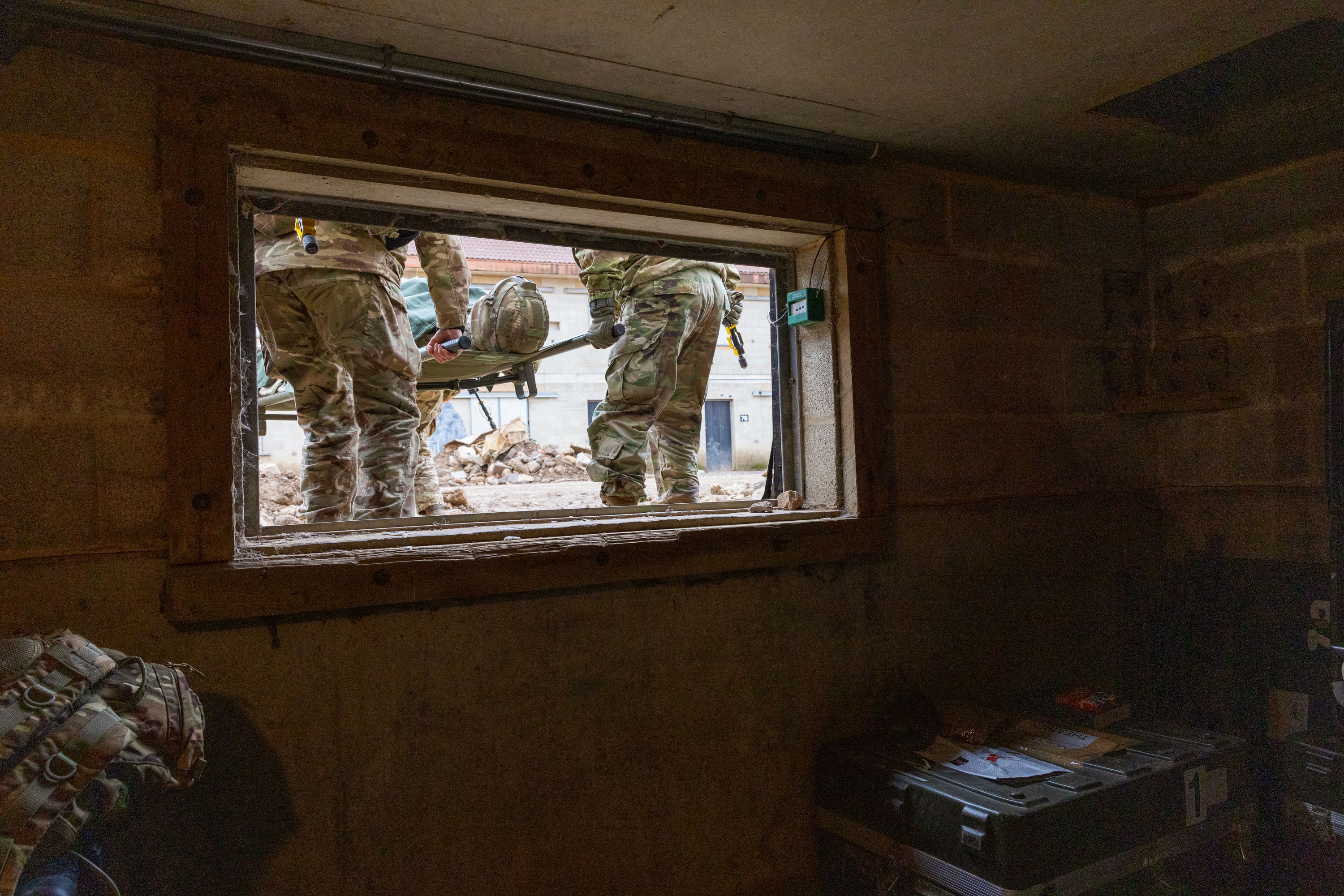 Two soldiers in camouflage uniforms carry a stretcher outside a building, viewed through a rectangular window.
