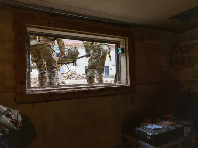 Two soldiers in camouflage uniforms carry a stretcher outside a building, viewed through a rectangular window.