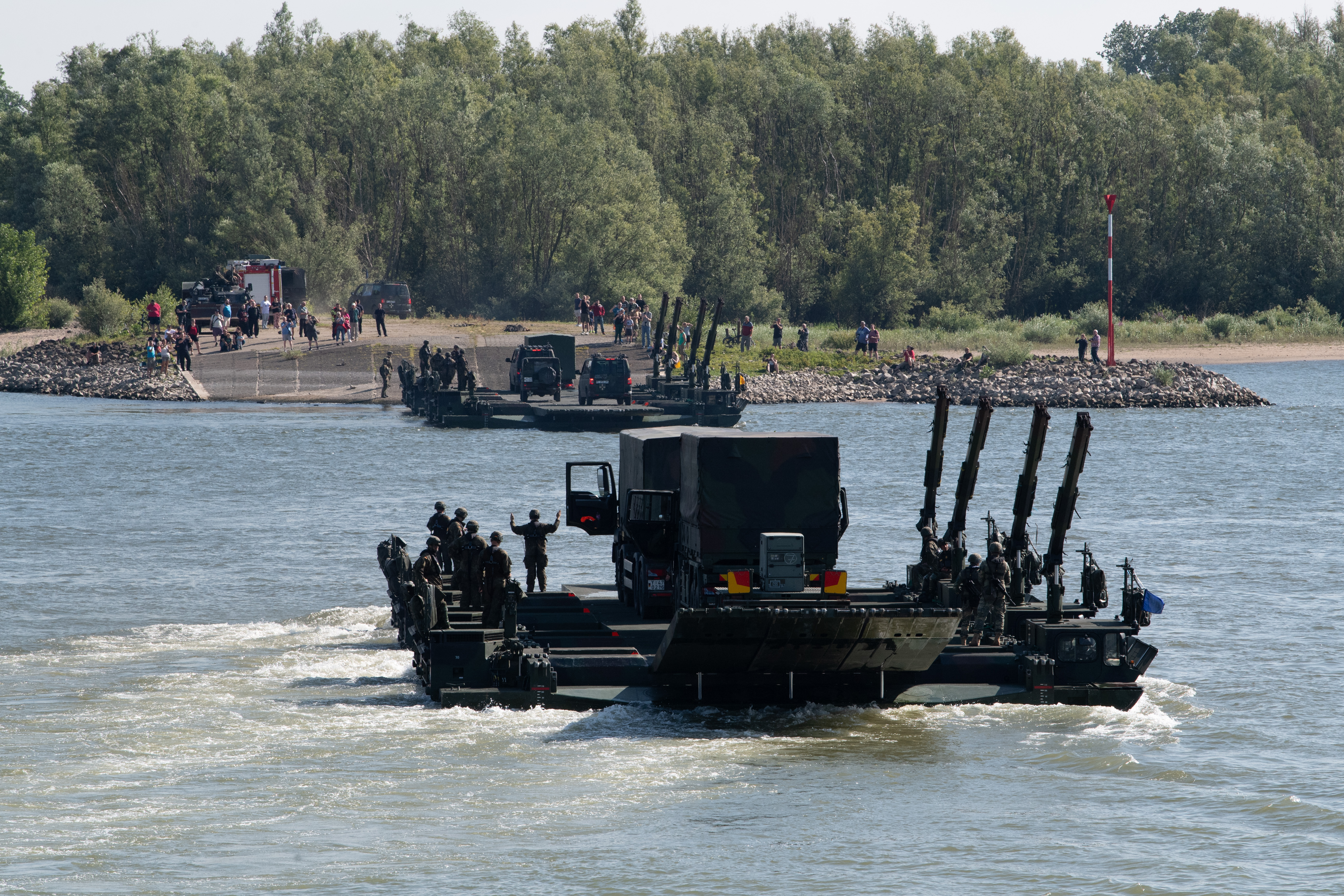 Military personnel and vehicles crossing a river with a wooded shoreline in the background.