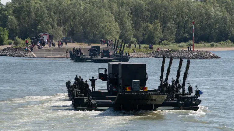 Military personnel and vehicles crossing a river with a wooded shoreline in the background.