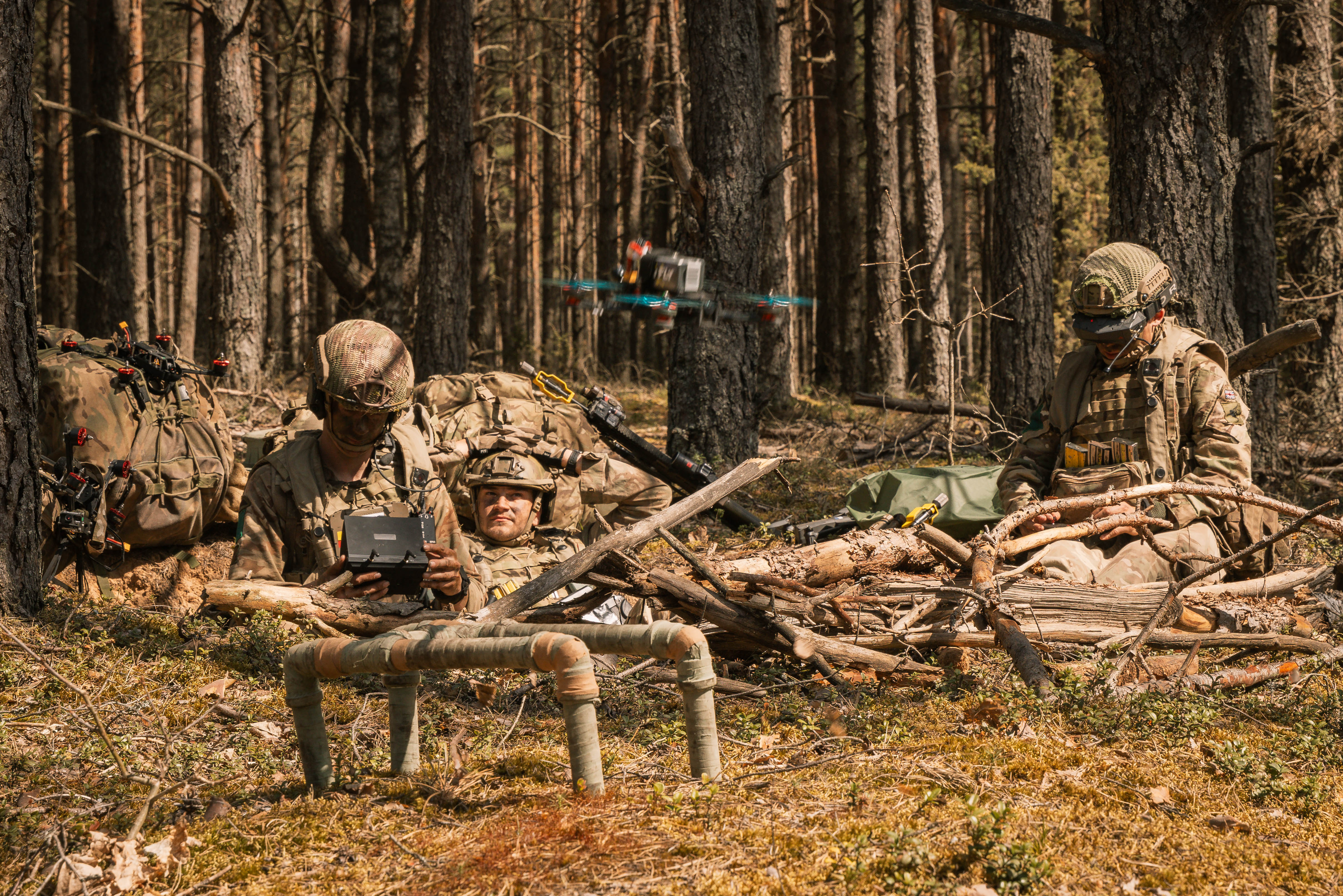 Soldiers in camouflage rest in a forest, operating a drone. They're surrounded by backpacks and branches, conveying a sense of focus and preparedness.