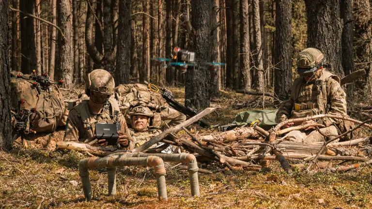 Soldiers in camouflage rest in a forest, operating a drone. They're surrounded by backpacks and branches, conveying a sense of focus and preparedness.