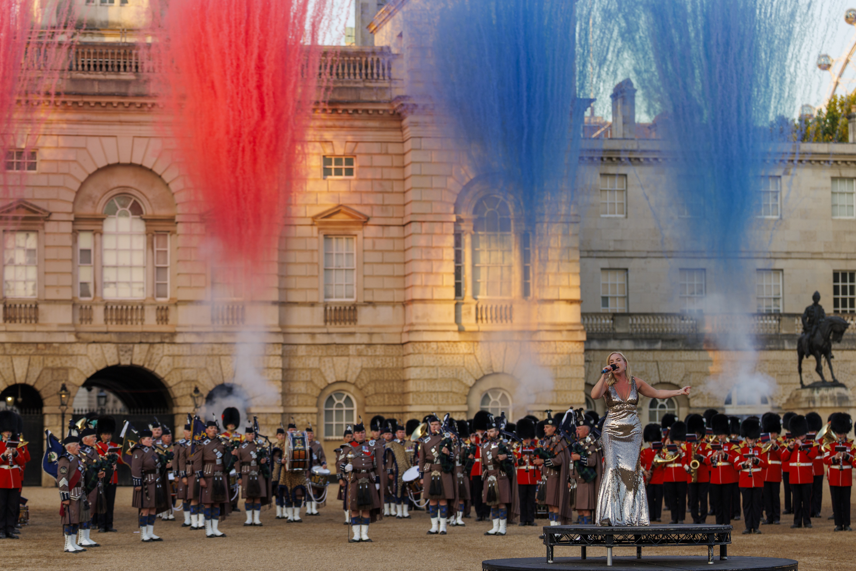 A performer in a silver outfit sings on a stage in front of the Band of the Scots Guards and massed pipers, who are dressed in traditional uniforms. Behind them, colorful plumes of red, white, and blue smoke rise into the sky at Horse Guards Parade in London.