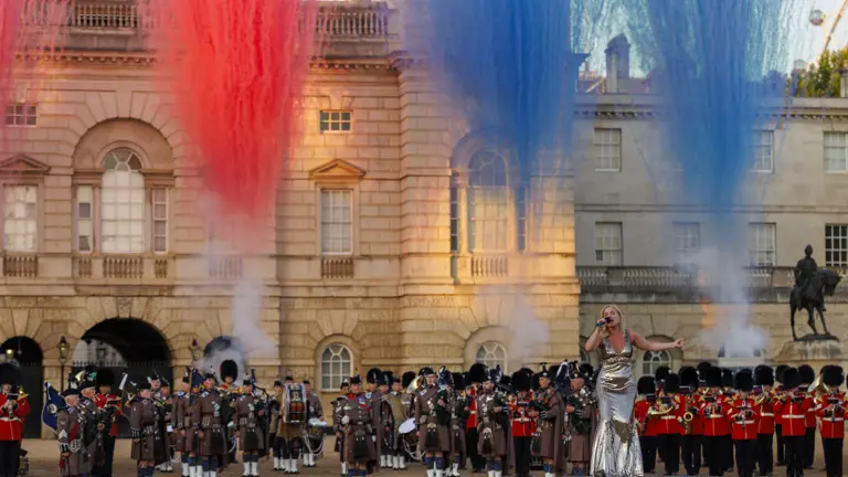 A performer in a silver outfit sings on a stage in front of the Band of the Scots Guards and massed pipers, who are dressed in traditional uniforms. Behind them, colorful plumes of red, white, and blue smoke rise into the sky at Horse Guards Parade in London.
