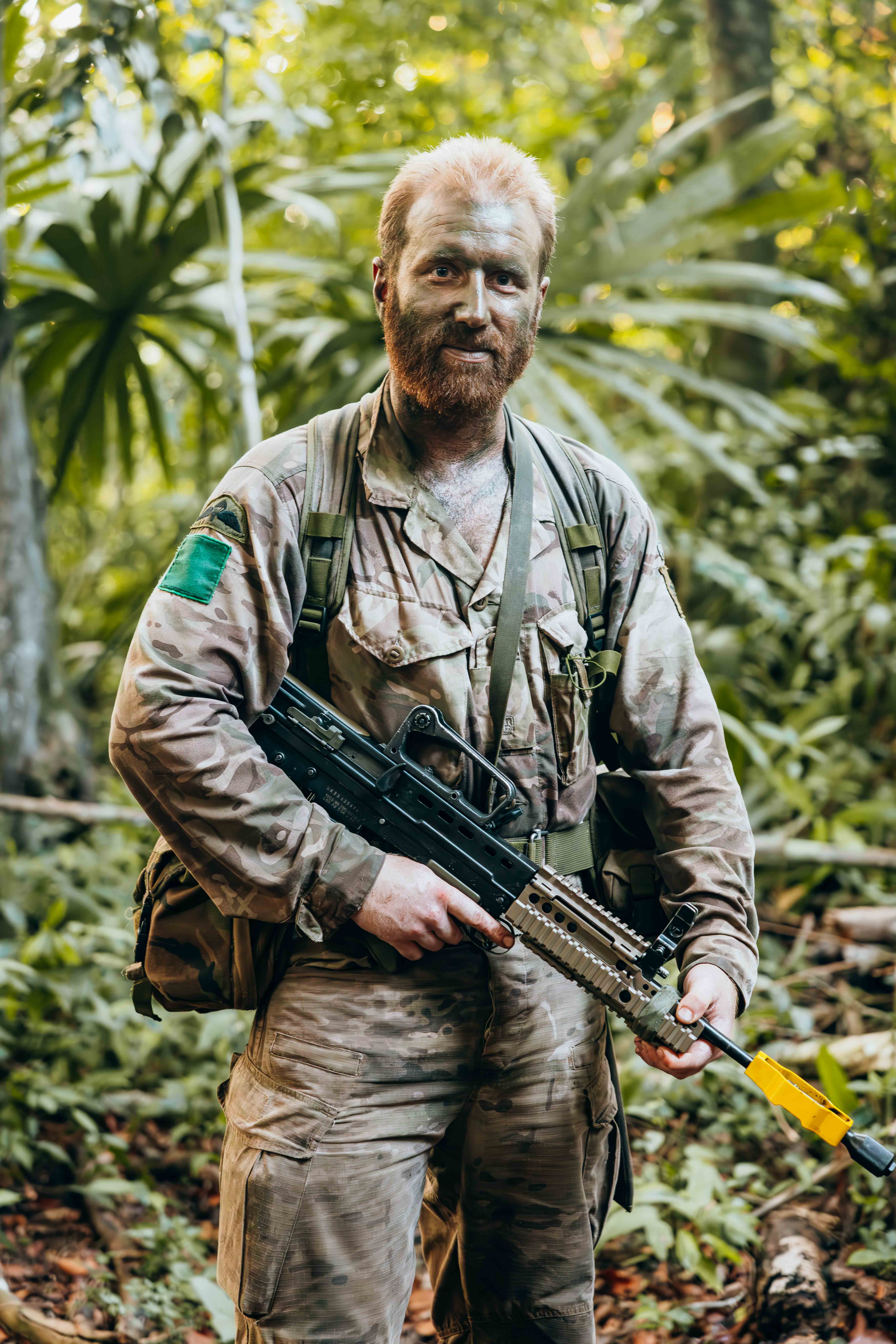 Soldier in full camouflage gear holding a rifle while standing in a dense jungle environment.