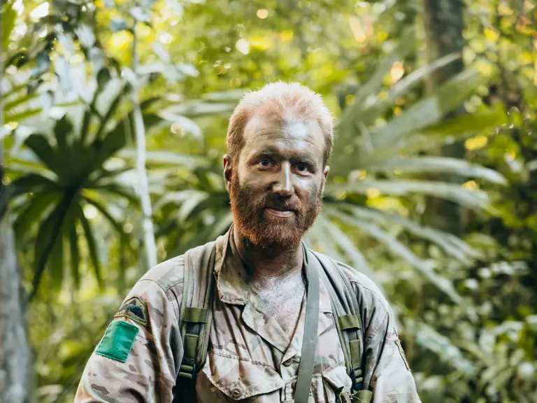 Soldier in full camouflage gear holding a rifle while standing in a dense jungle environment.