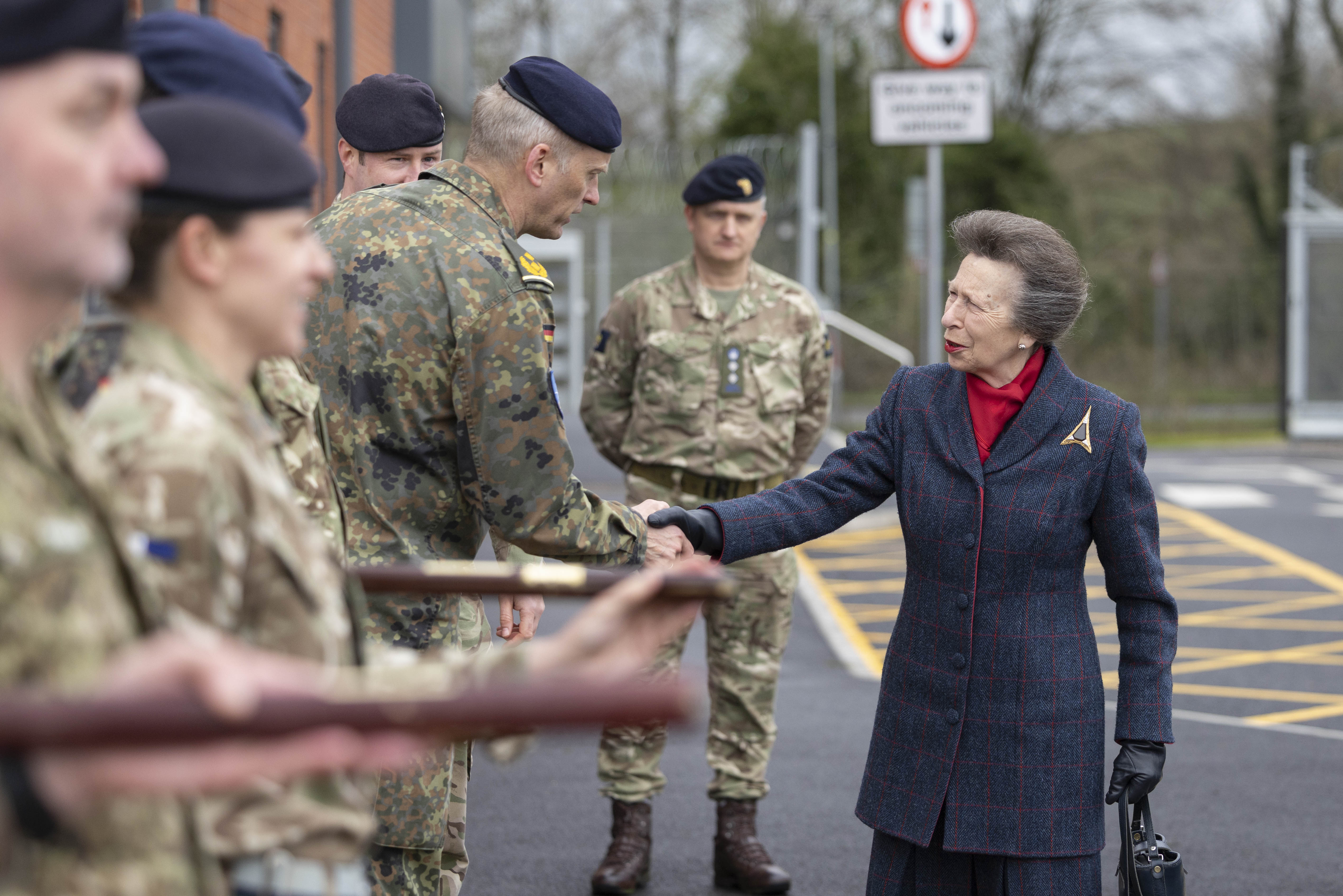 A person wearing a red plaid coat stands among several uniformed military personnel. They are engaged in an outdoor ceremonial event, with the military personnel holding rifles.