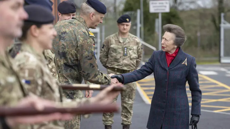 A person wearing a red plaid coat stands among several uniformed military personnel. They are engaged in an outdoor ceremonial event, with the military personnel holding rifles.