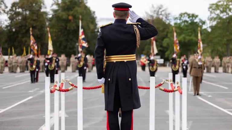 A person in a formal military uniform stands on a platform, saluting a group of soldiers holding flags. The soldiers are arranged in formation across an open area. The scene is framed by trees in the background.