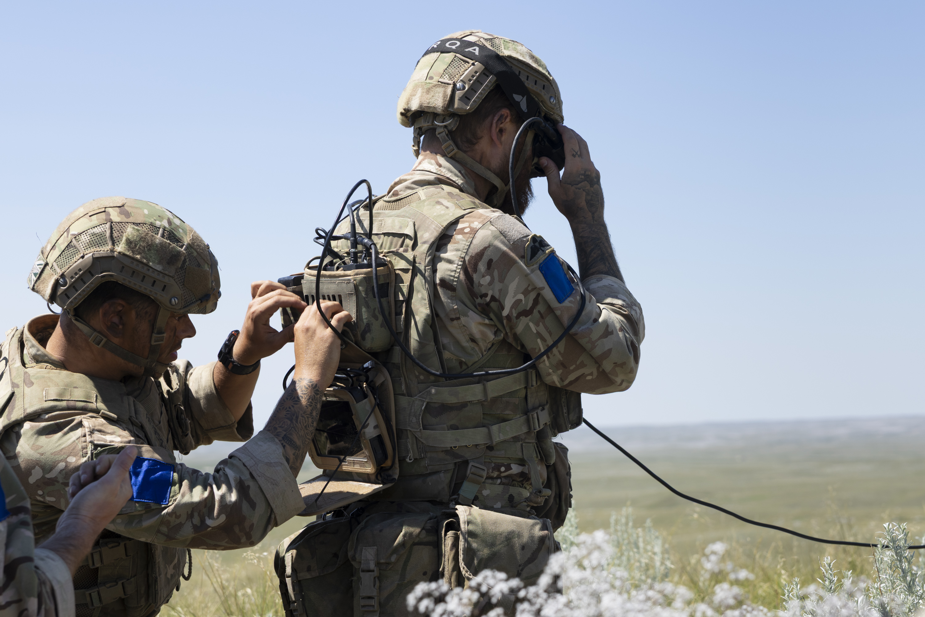 Two soldiers in camouflage gear and helmets operate communication equipment in an open field. One adjusts a backpack radio, while the other uses a handheld device. The scene suggests a military exercise or communication task.