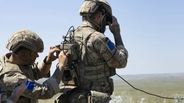 Two soldiers in camouflage gear and helmets operate communication equipment in an open field. One adjusts a backpack radio, while the other uses a handheld device. The scene suggests a military exercise or communication task.