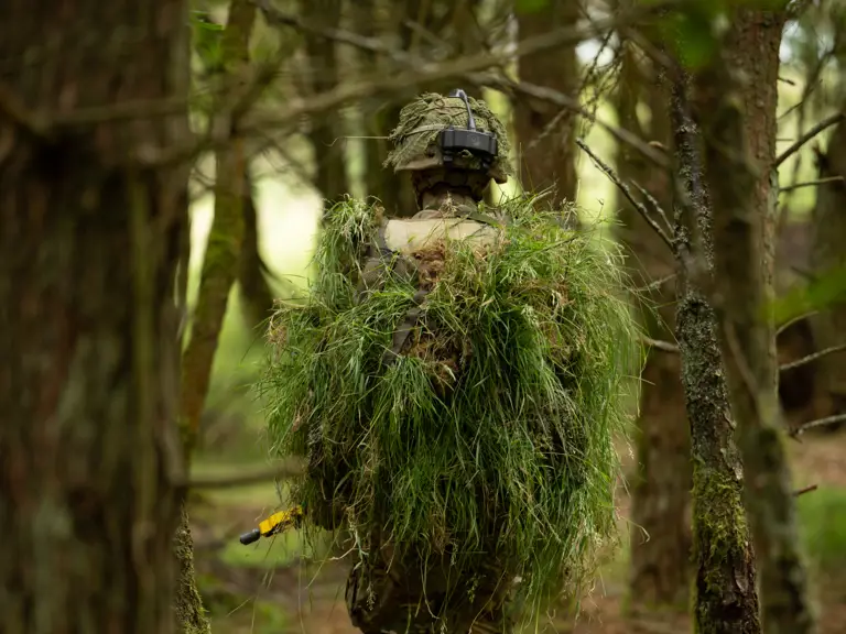A person wearing camouflage and a helmet walks through a forest, carrying a large bundle of green foliage on their back.