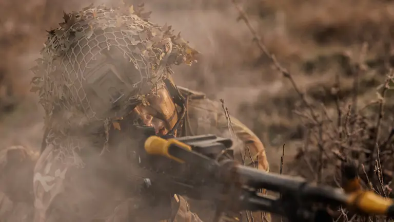 Soldier seen on exercise wearing camouflage uniform, camouflage cream and a helmet shooting a machine gun.