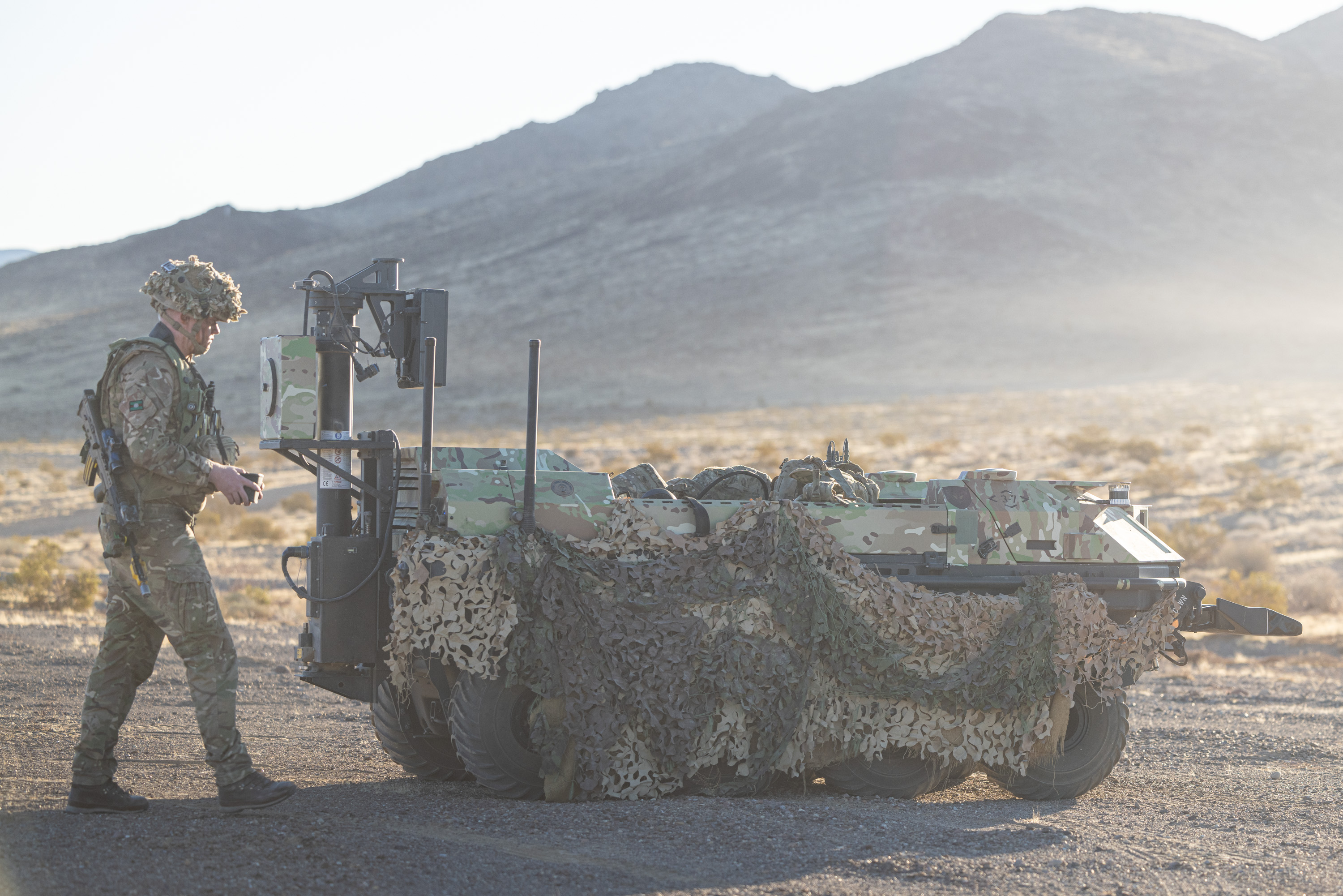 A soldier walks behind a green vehicle covered in camouflage nets.