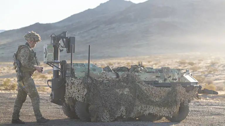 A soldier walks behind a green vehicle covered in camouflage nets.