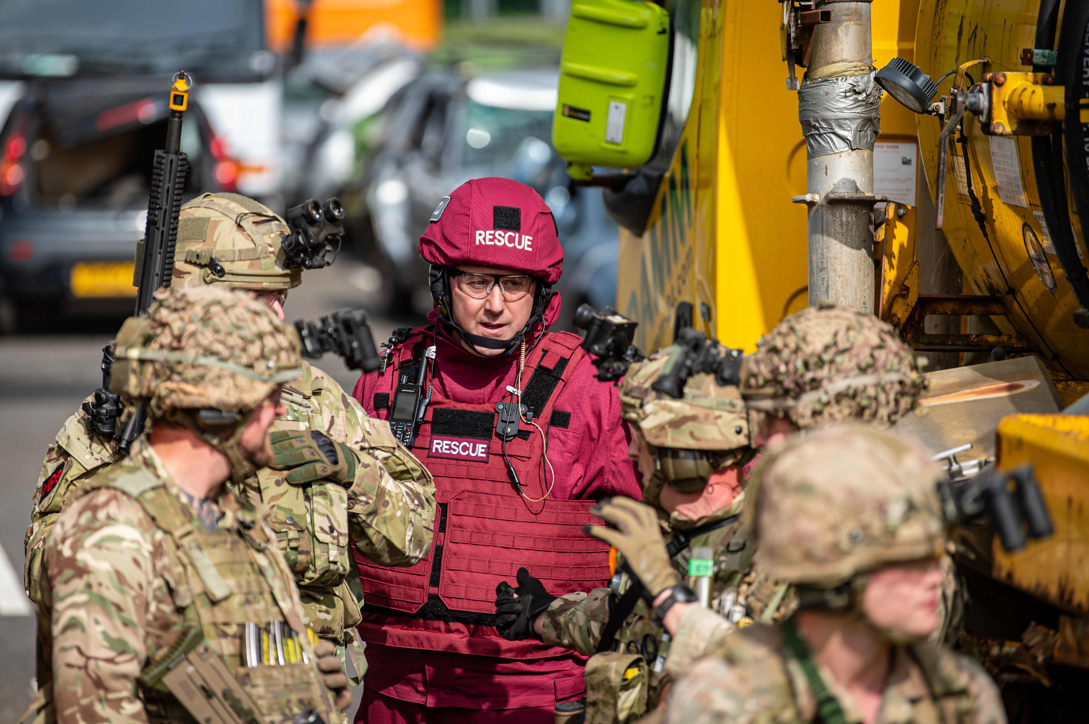 A group of individuals in military and rescue uniforms stands next to a large yellow vehicle labeled 