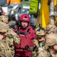 A group of individuals in military and rescue uniforms stands next to a large yellow vehicle labeled