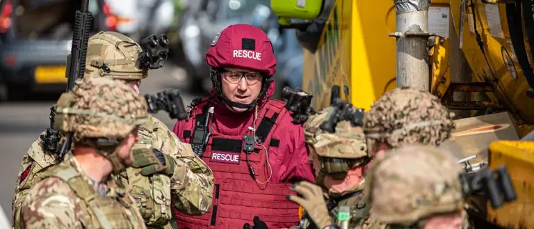 A group of individuals in military and rescue uniforms stands next to a large yellow vehicle labeled