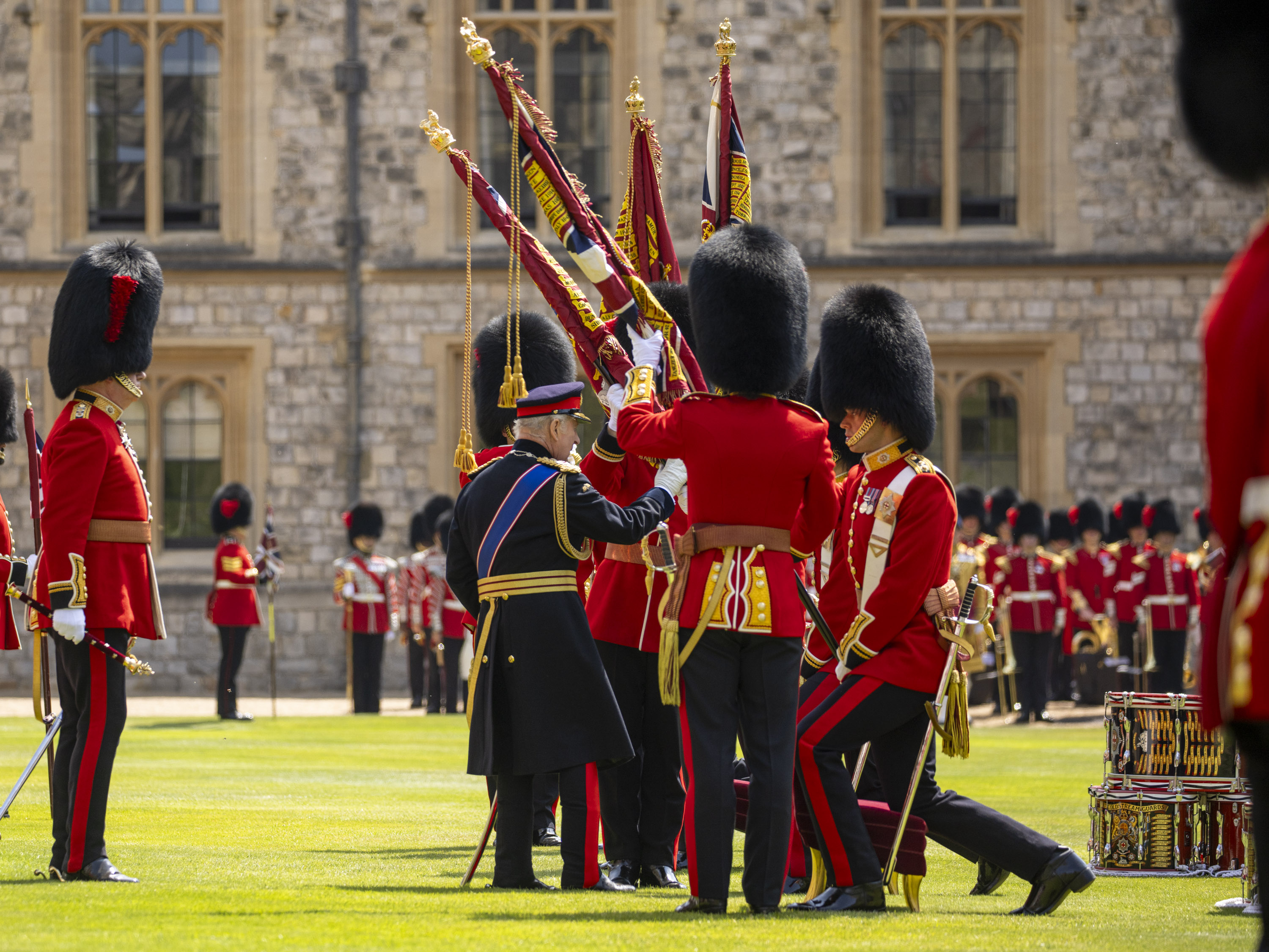 British guards in traditional red uniforms and bearskin hats holding ceremonial flags on a lawn.