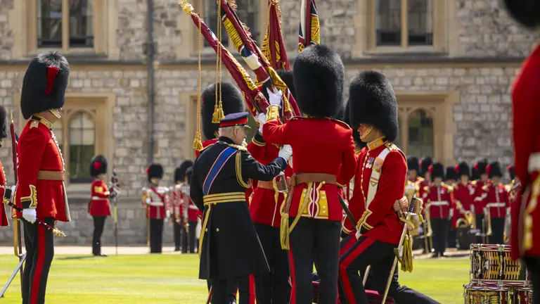British guards in traditional red uniforms and bearskin hats holding ceremonial flags on a lawn.