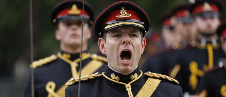 A person in a dress uniform, adorned with multiple medals on the chest, is leading a group of similarly dressed personnel