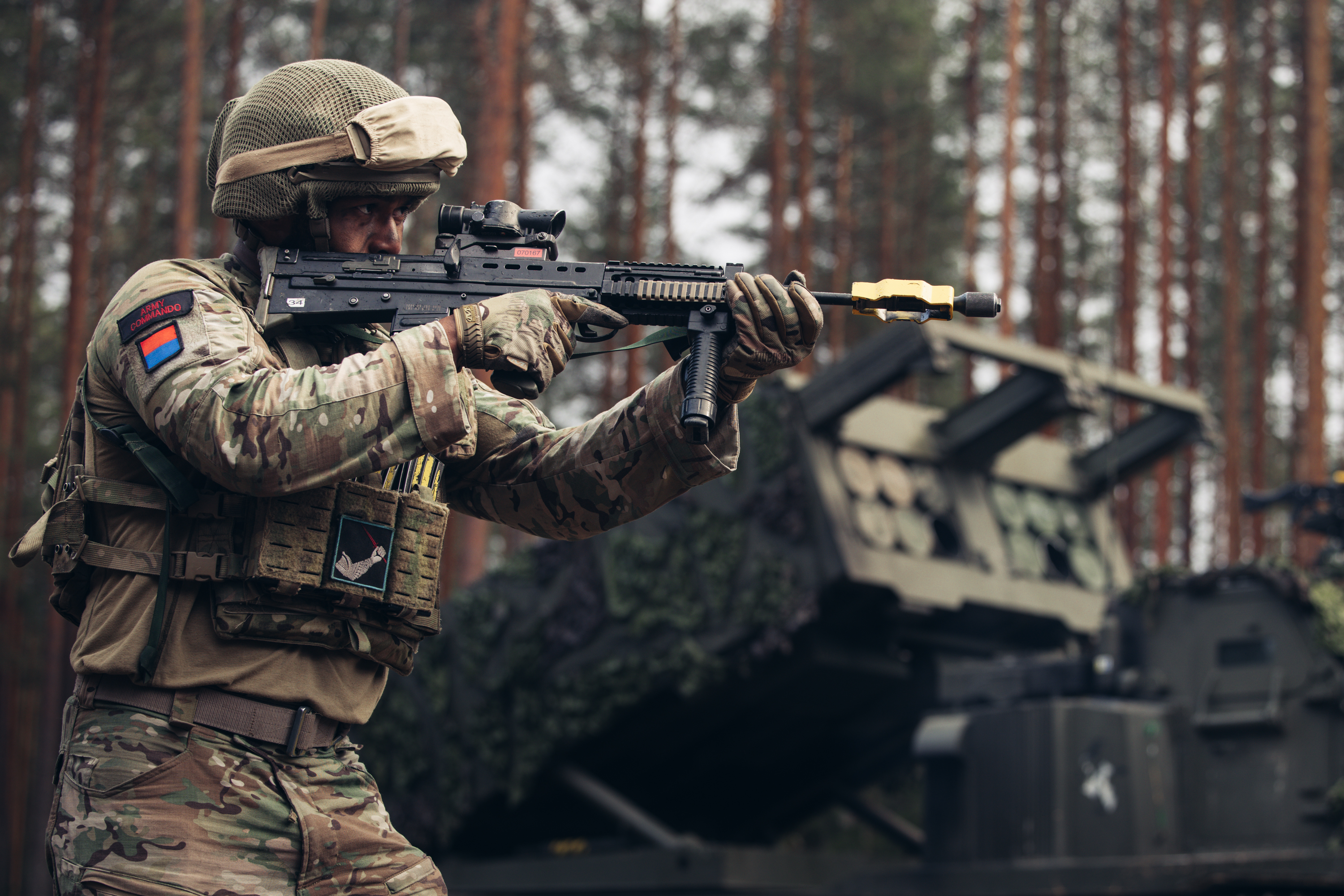 Soldier in camouflage gear aiming a rifle in a forested area with military equipment in the background.