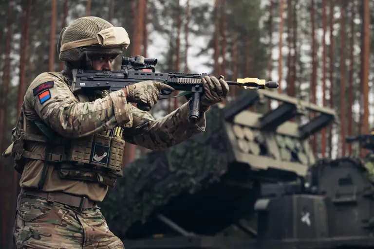 Soldier in camouflage gear aiming a rifle in a forested area with military equipment in the background.