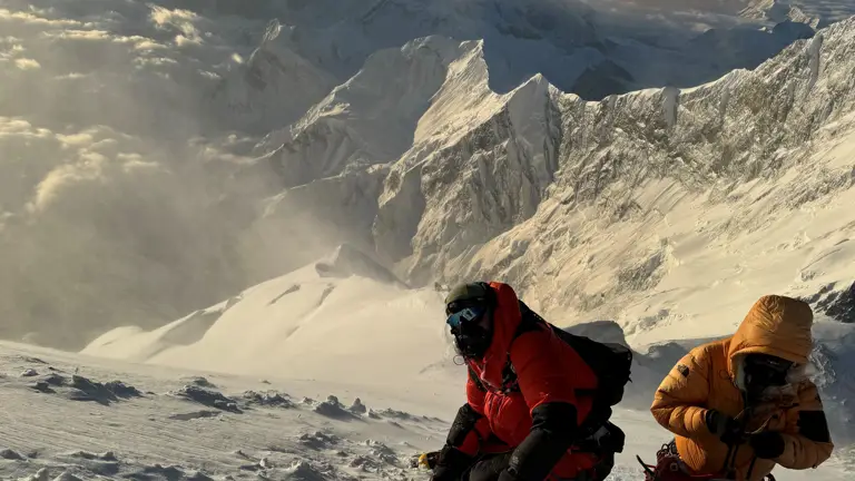 Two individuals in large coats and altitude masks are pictured against the backdrop of snowy mountains.
