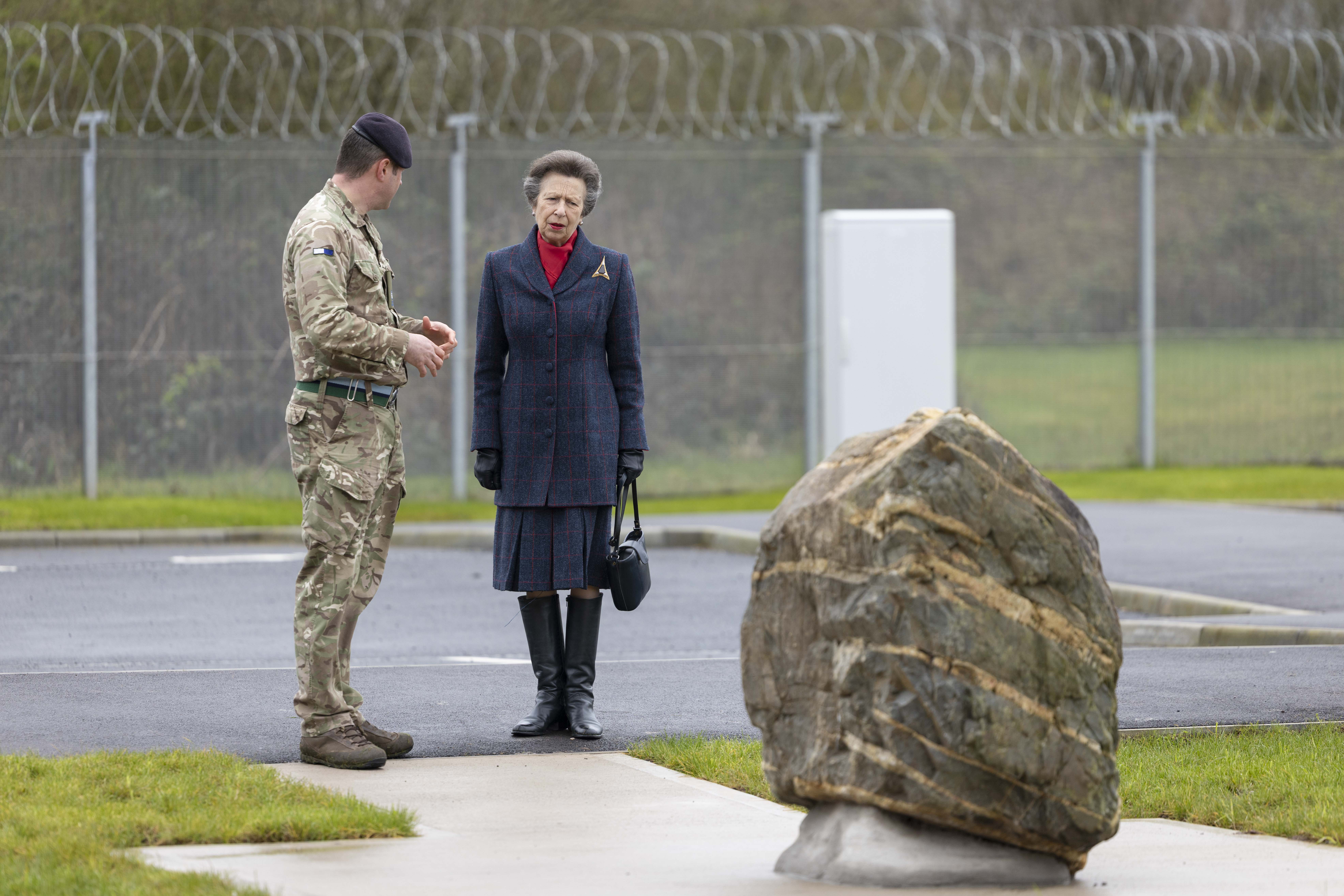 A woman in a dark coat and boots stands next to a soldier in camouflage, conversing near a large rock. The background features a barbed wire fence.