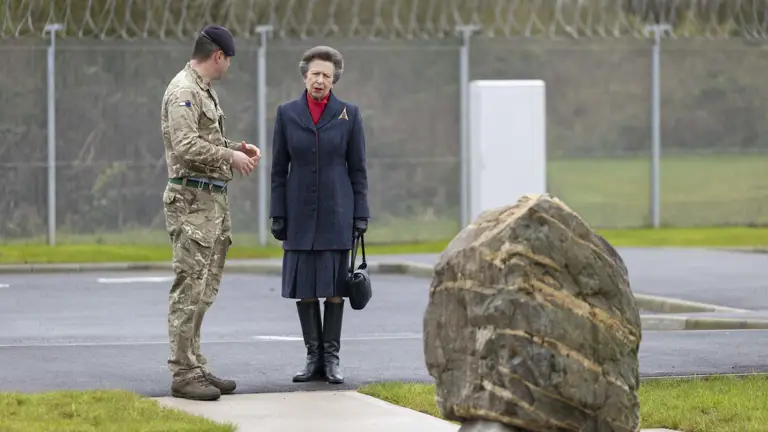 A woman in a dark coat and boots stands next to a soldier in camouflage, conversing near a large rock. The background features a barbed wire fence.