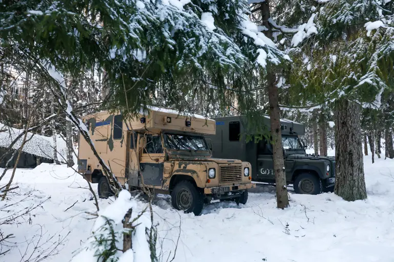 Two Land Rover vehicles parked in a snowy forest. One vehicle is tan, and the other is dark green. 