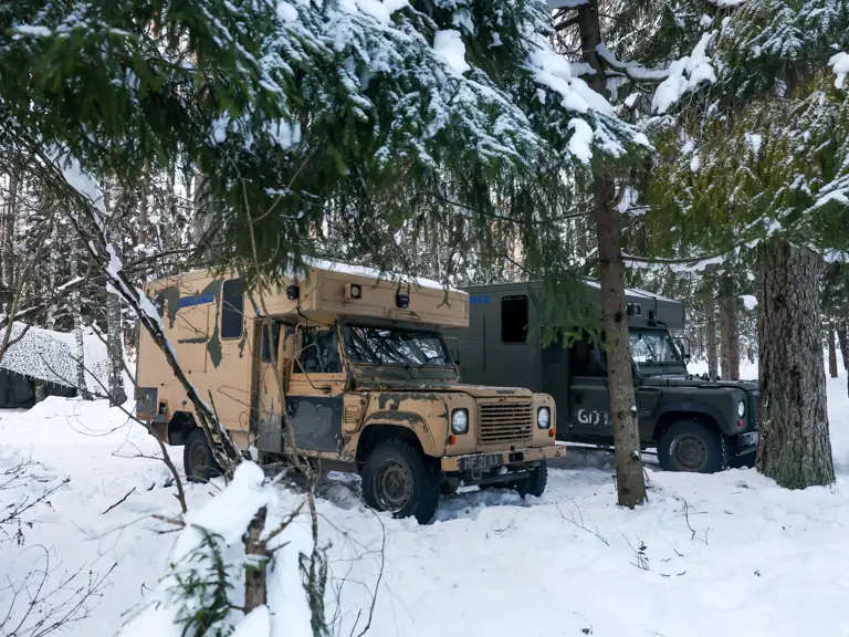Two Land Rover vehicles parked in a snowy forest. One vehicle is tan, and the other is dark green.