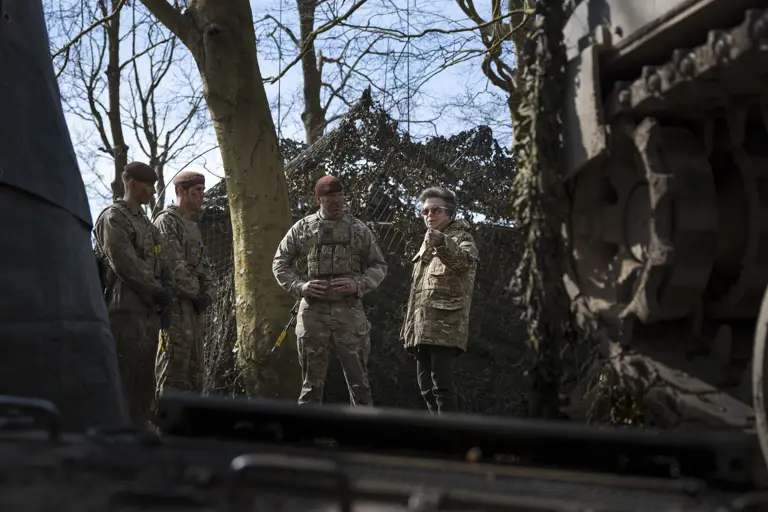 A group of military personnel in camouflage stand near a large piece of equipment, possibly a vehicle, in a wooded area.