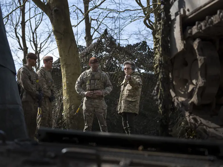 A group of military personnel in camouflage stand near a large piece of equipment, possibly a vehicle, in a wooded area.