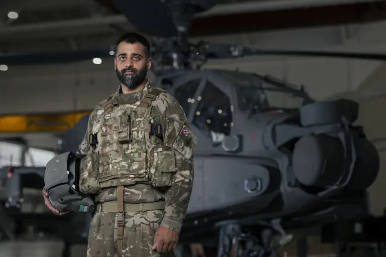 Soldier wearing camouflage uniform holding a helmet under one arm stands inside an aircraft hanger in front of an apache helicopter in the background.