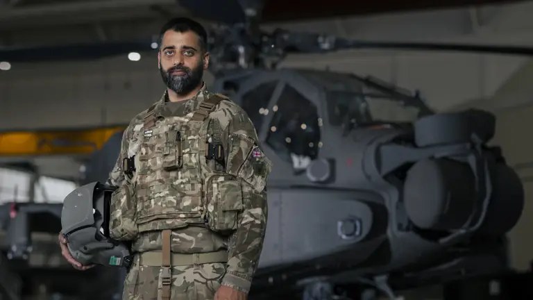 Soldier wearing camouflage uniform holding a helmet under one arm stands inside an aircraft hanger in front of an apache helicopter in the background.