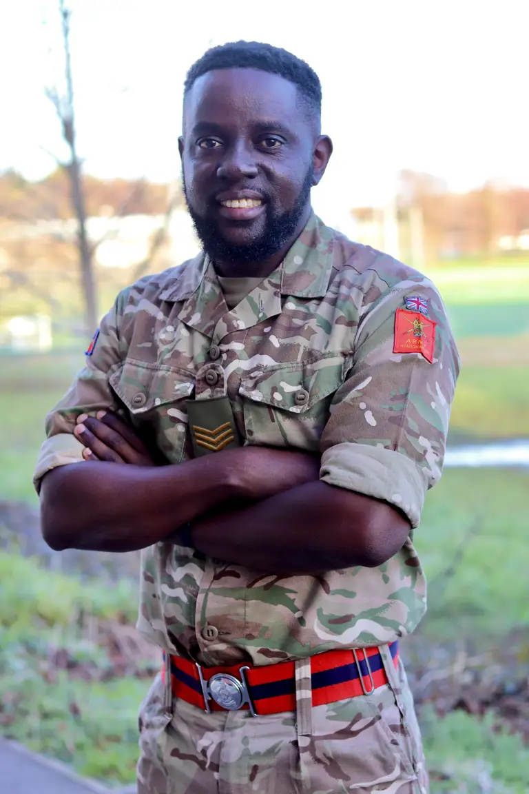 British Army soldier in camouflage uniform with sergeant rank insignia and red-striped belt standing outdoors.