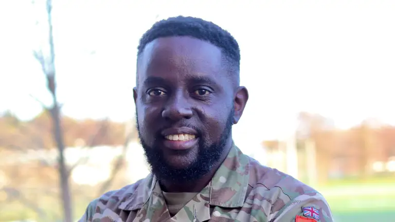 British Army soldier in camouflage uniform with sergeant rank insignia and red-striped belt standing outdoors.